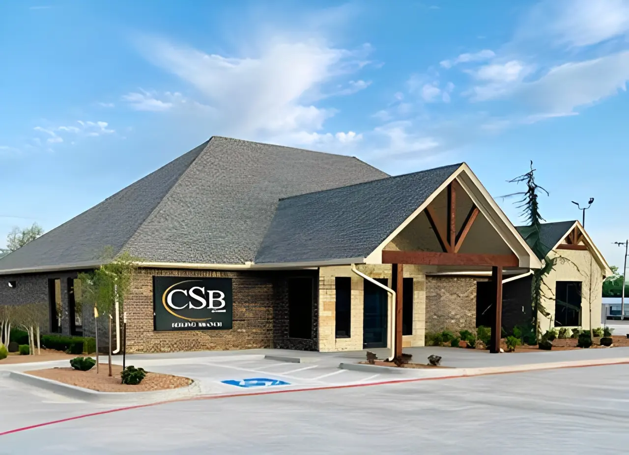 A modern brick and stone building with a dark gray roof, labeled "CSB", set against a blue sky. The entrance features wooden beams, and a wheelchair-accessible parking space is visible in front.