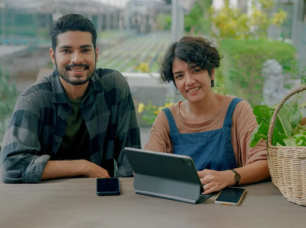 Two people sit at a table with a tablet, smartphone, and a basket of plants. They are looking at the camera and smiling, with greenery in the background.