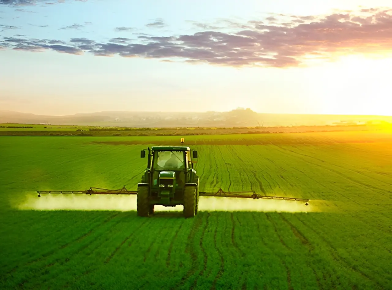 A tractor sprays crops in a vast green field under a vibrant sunset sky.